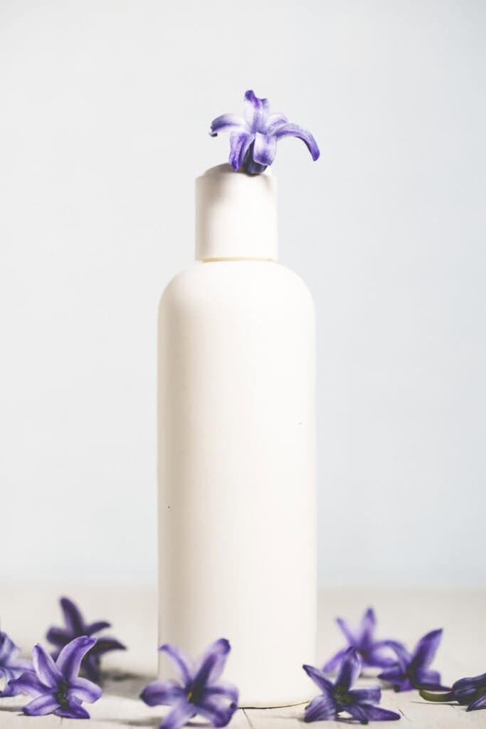 A blank shampoo bottle with a purple flower blooming out of the lid with more purple flowers surrounding the base.