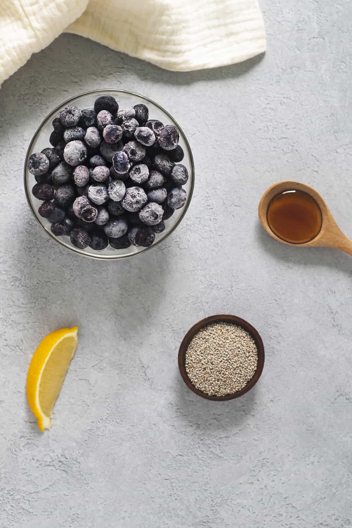 A bowl of frozen blueberries, a slice of fresh lemon, a bowl of chia seeds and a spoonful of 100% pure maple syrup.