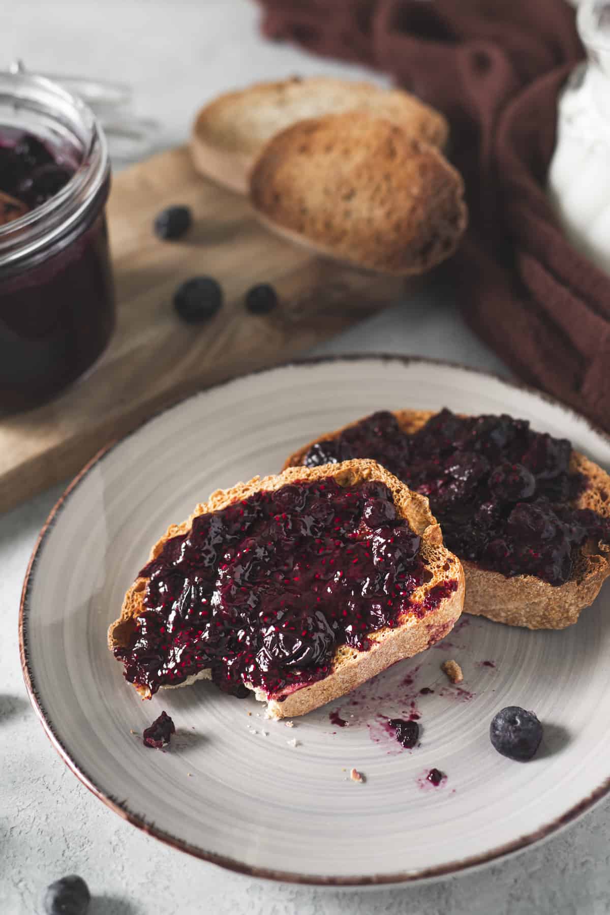 A plate of semi-eaten blueberry jam toast served beside a full jam jar, extra slices of toast and spilled whole blueberries.