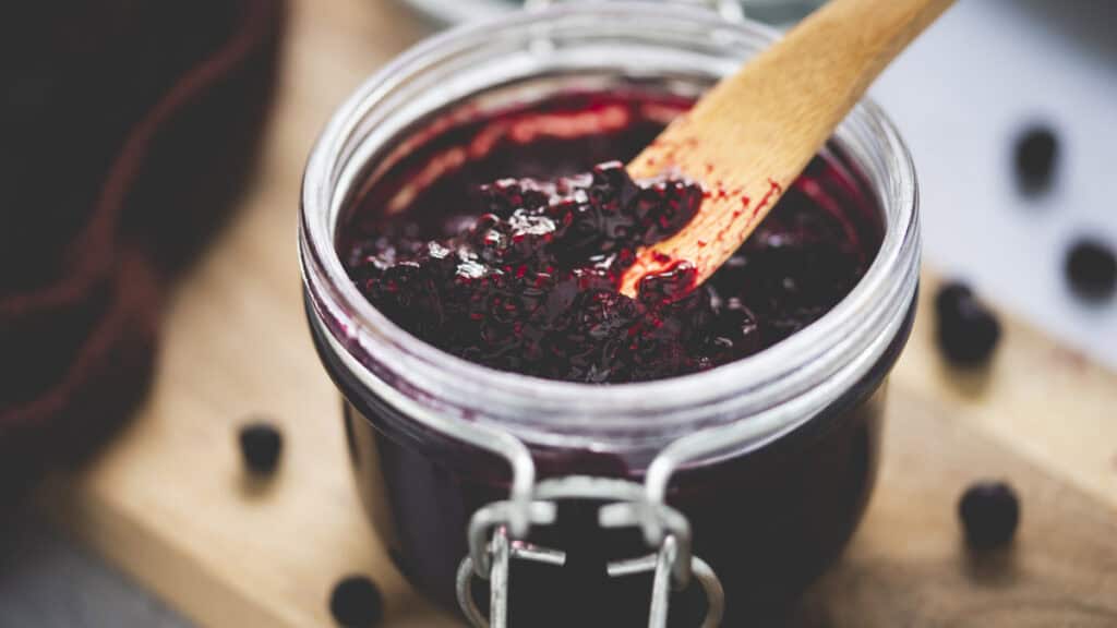 Surrounded by spilled whole blueberries and a clove-brown cloth, an open jar of blueberry chia jam sits atop a wooden serving board with a wooden butter knife.