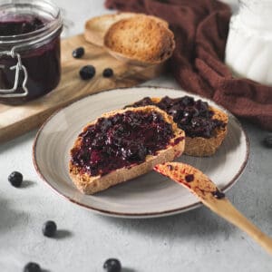 A plate of toast with freshly spread blueberry jam served on a breakfast table with spilled blueberries, a milk creamer, an open jam jar and a clove-brown cloth.