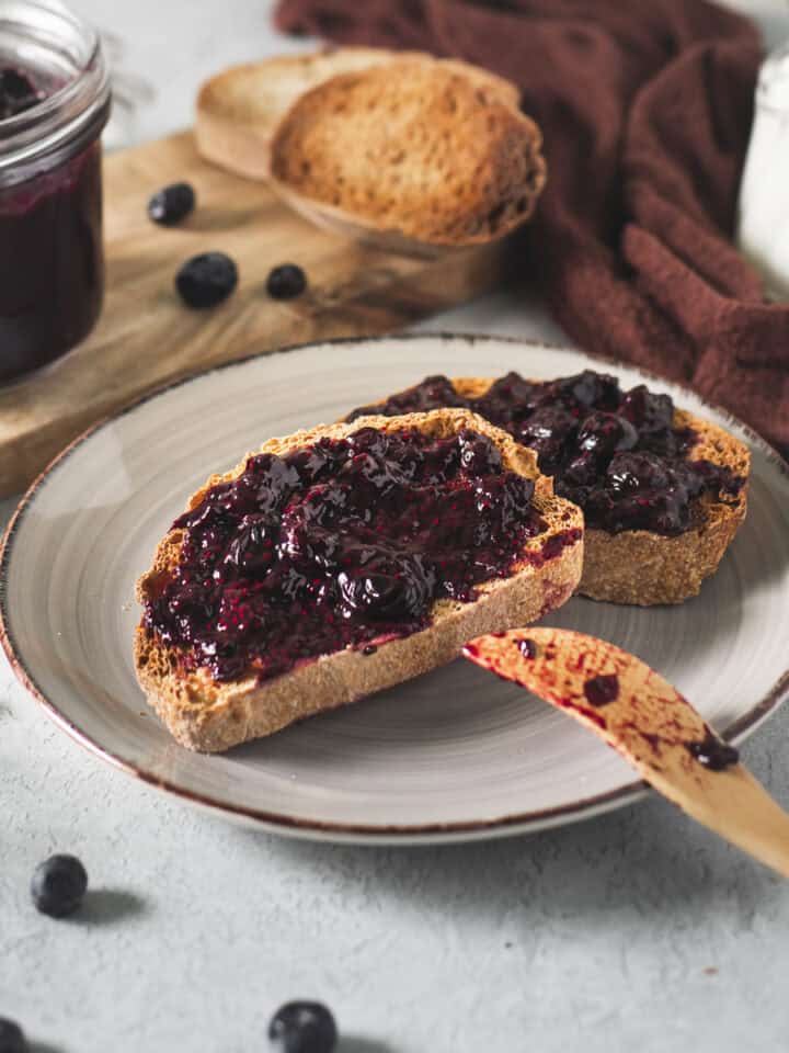 A plate of toast with freshly spread blueberry jam served on a breakfast table with spilled blueberries, a milk creamer, an open jam jar and a clove-brown cloth.