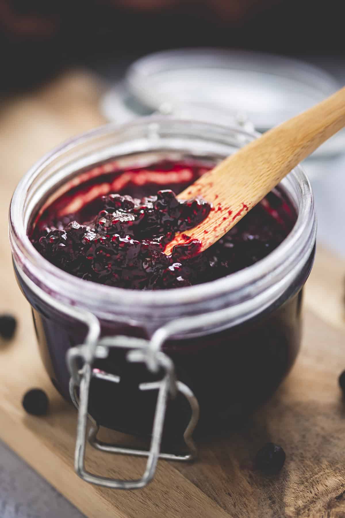 Surrounded by spilled whole blueberries and a clove-brown cloth, an open jar of blueberry chia jam sits atop a wooden serving board with a wooden butter knife.