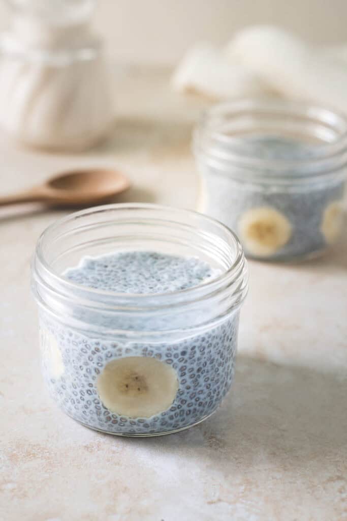 Two jars of blue chia pudding with sliced bananas garnished around the sides. A wooden spoon and jug of almond milk sitting in the background.