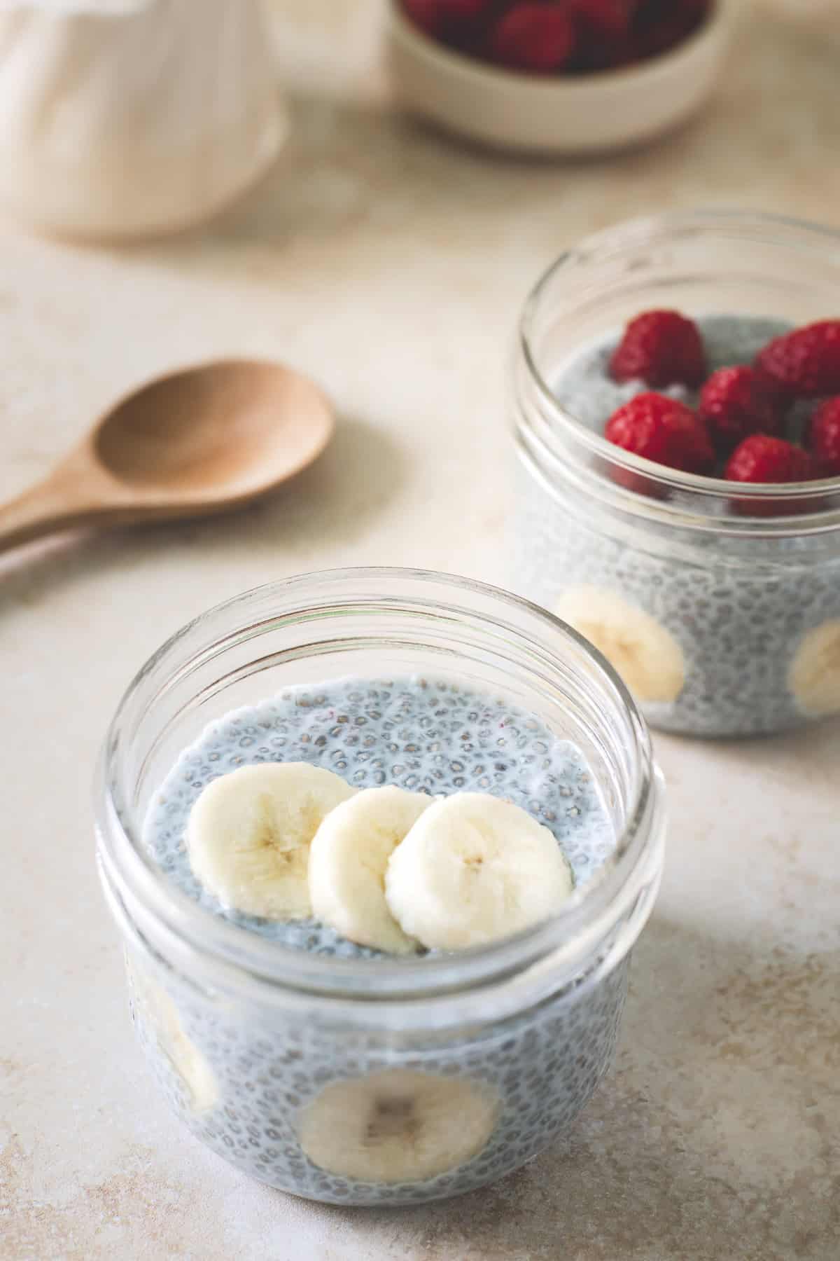 Two jars of blue chia pudding, one topped with raspberries and one with sliced banana. A bowl of fresh berries placed behind with a wooden spoon and a jug of milk.