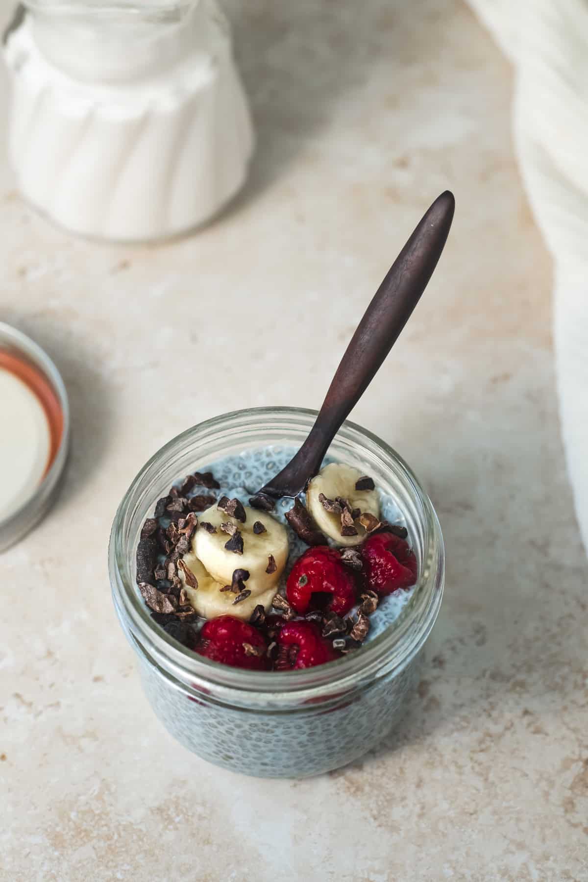 A jar of blue chia seed pudding garnished with sliced banana, raspberries and cacao nibs.