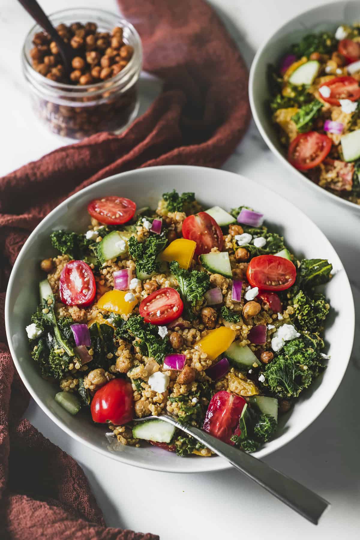 Two bowls of quinoa salad full of fresh veggies, a jar roasted chickpeas and a burnt orange table runner.