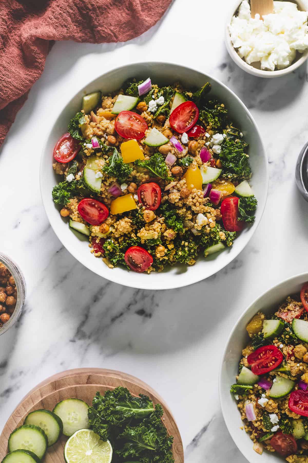 Two large bowls of Quinoa Salad served on a table with fresh cucumber, dressing roasted chickpeas and chèvre.