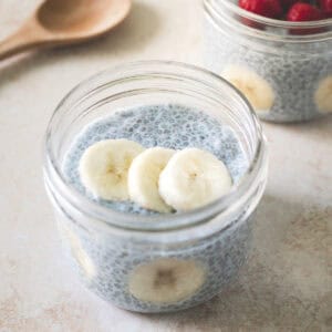 Two jars of blue chia pudding topped with raspberries and bananas beside a wooden spoon.