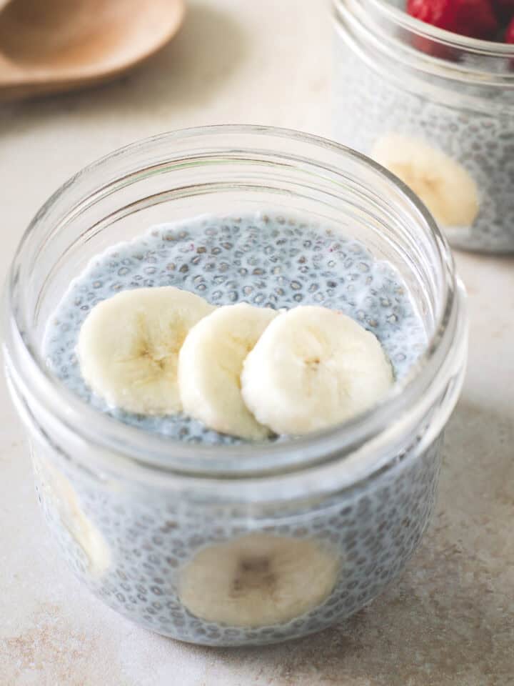 Two jars of blue chia pudding topped with raspberries and bananas beside a wooden spoon.