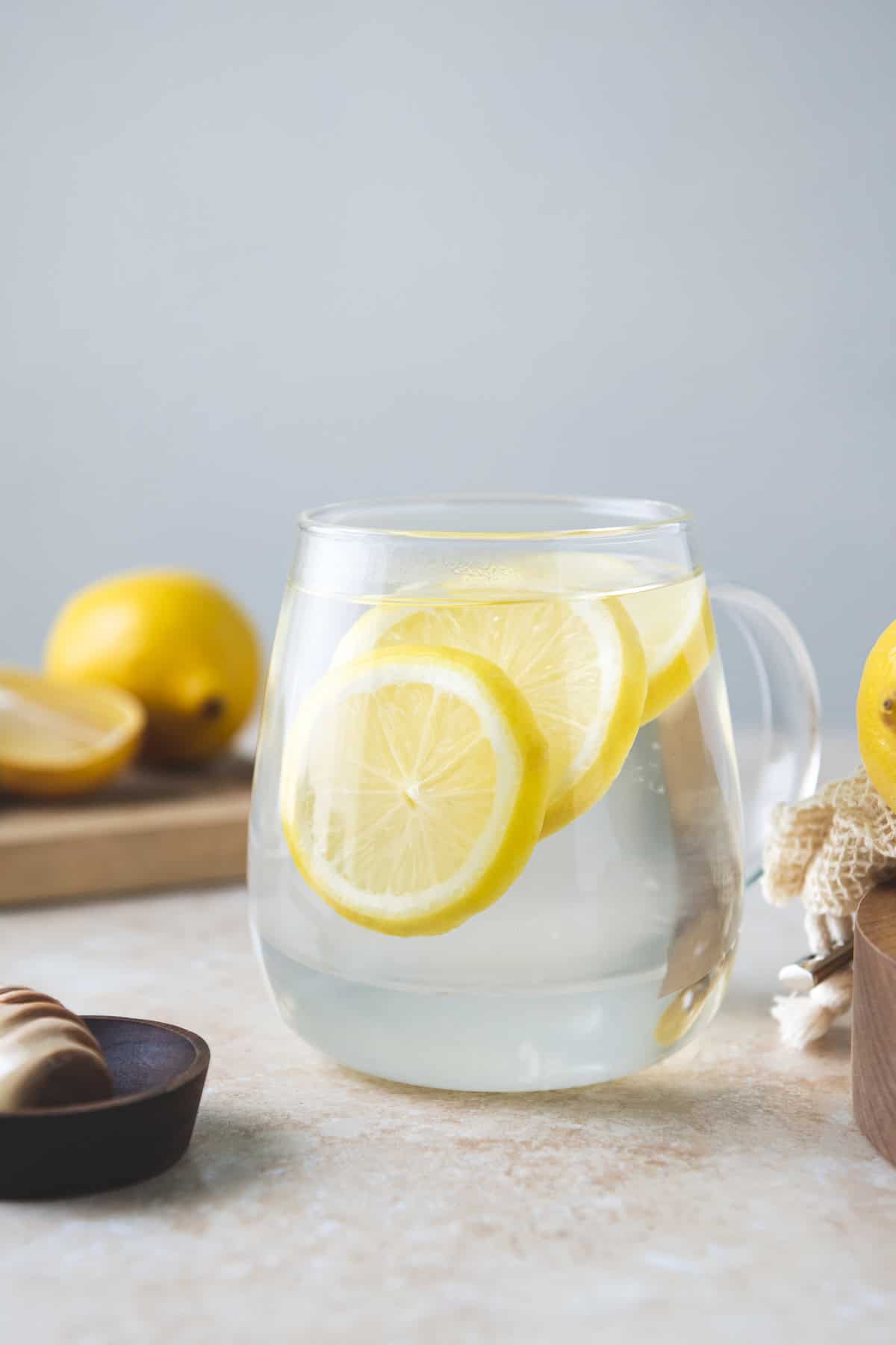 Three lemon slices floating in a mug of water beside a honey dipper and cutting board with whole and halved lemons.