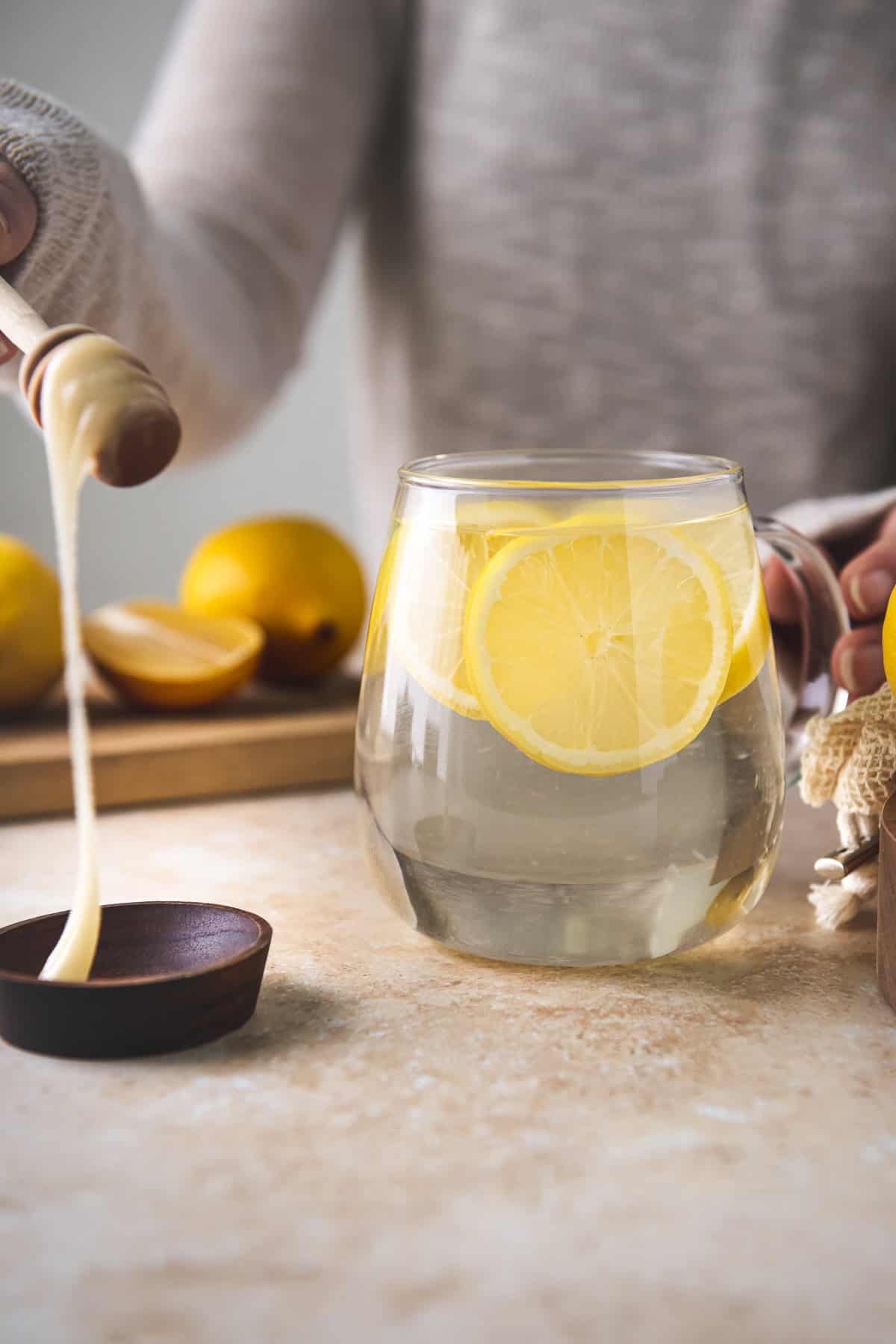 Honey dripping off a dipper into a bowl beside a mug of lemon water.