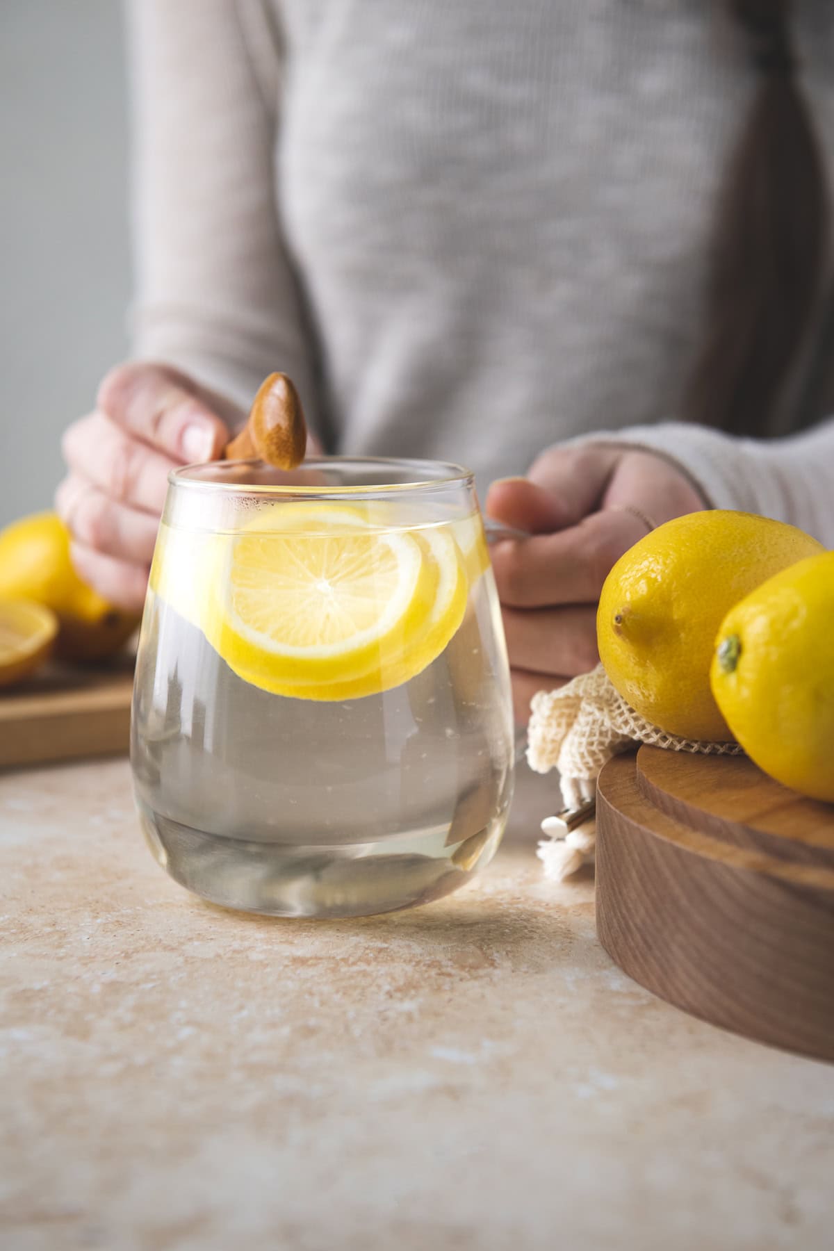 A dark-haired woman tapping a wooden spoon on a glass mug filled with sliced lemon and hot water.