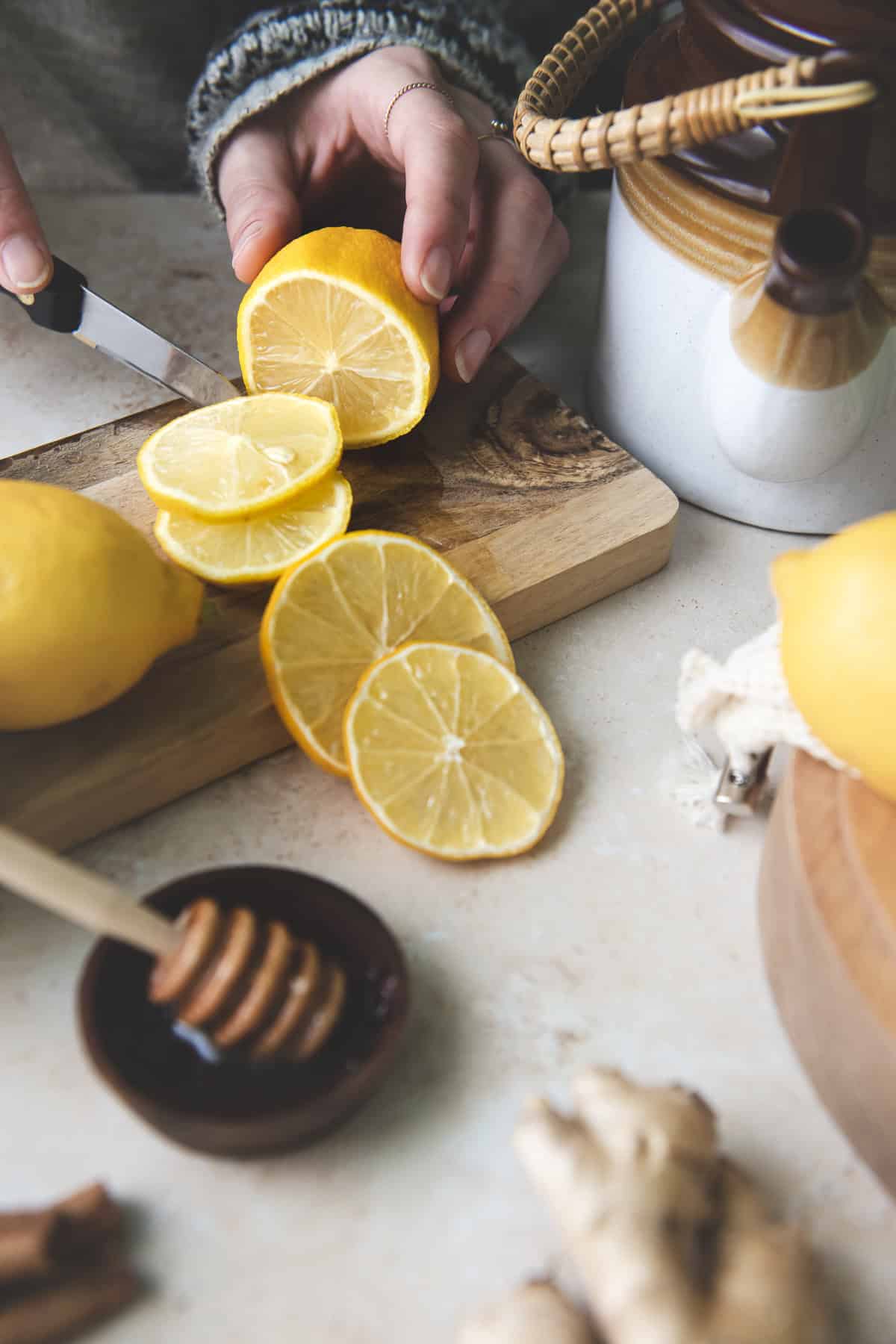 A lemon being sliced into rounds on a cutting board surrounded by a ceramic tea pot, a honey dipper and more whole lemons.